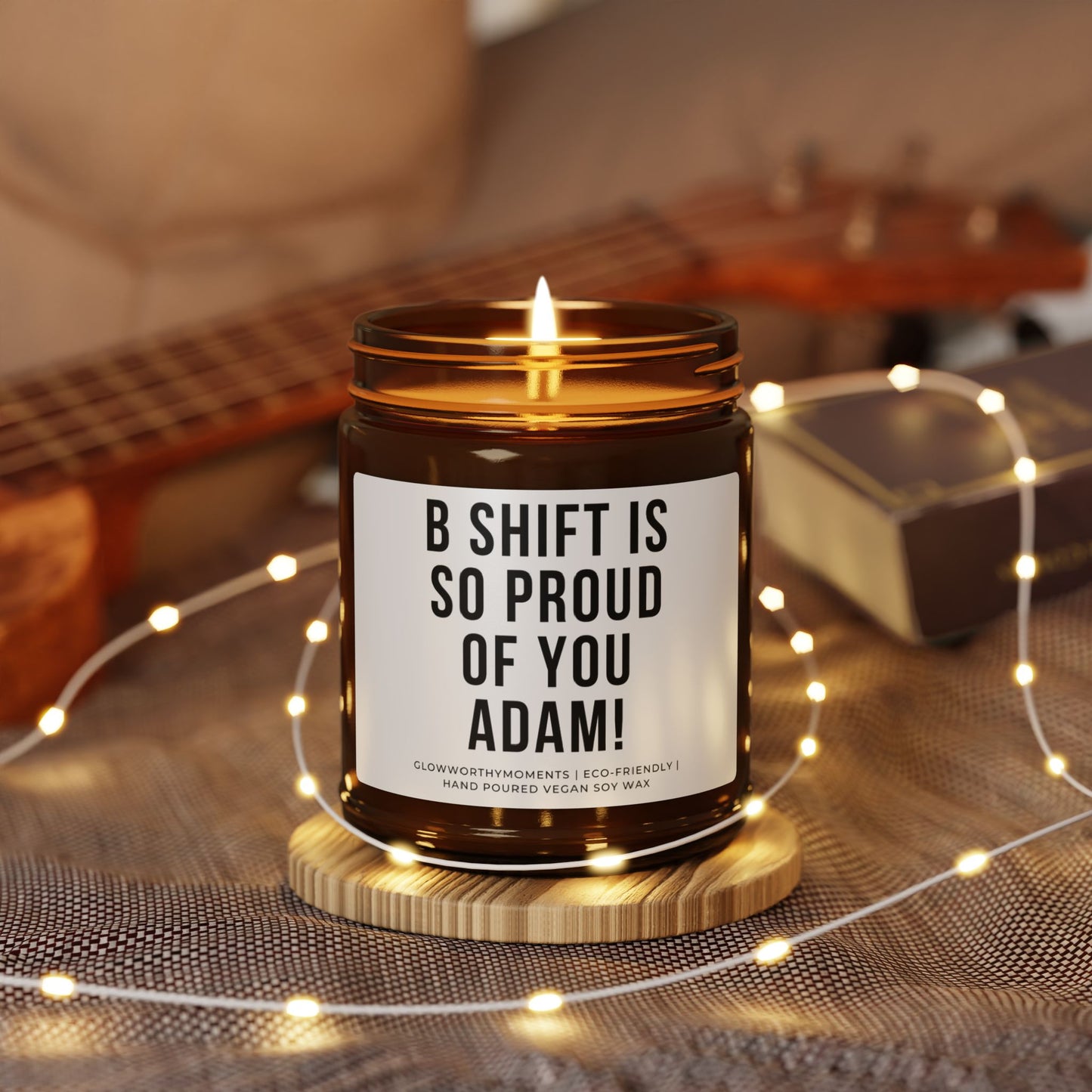 Candle in a jar with a personalized message on a wooden coaster surrounded by string lights and a guitar in the background.