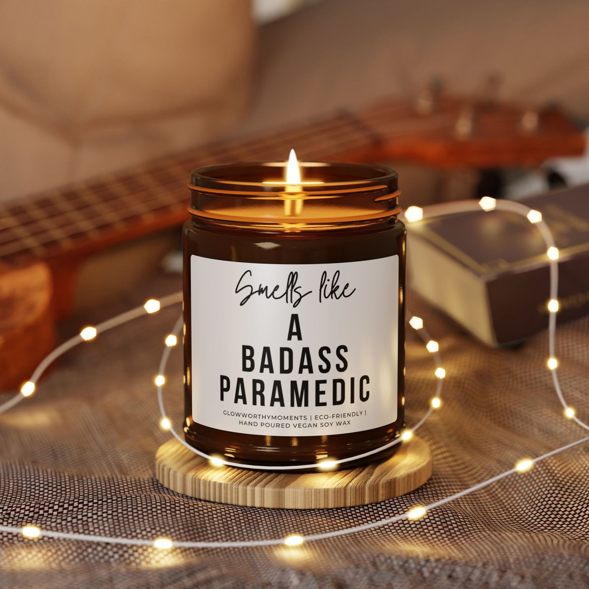 Candle in a jar with a label reading 'Smells like a badass paramedic' on a wooden coaster with fairy lights and a guitar in the background.