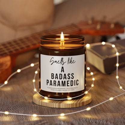 Candle in a jar with a label reading 'Smells like a badass paramedic' on a wooden coaster with fairy lights and a guitar in the background.