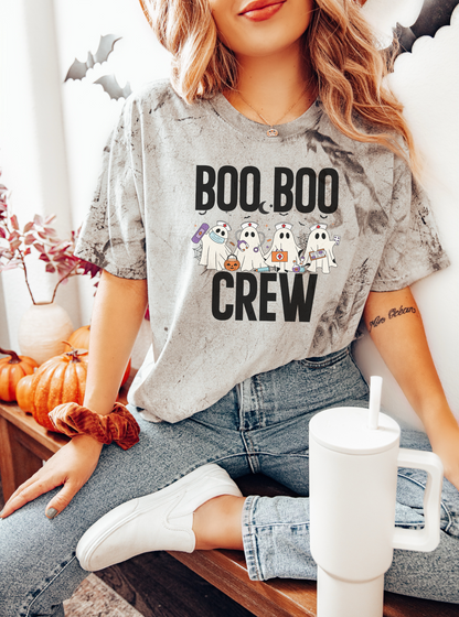 Person wearing a 'BOO BOO CREW' shirt with ghost graphics, sitting on a couch with pumpkins and a mug.