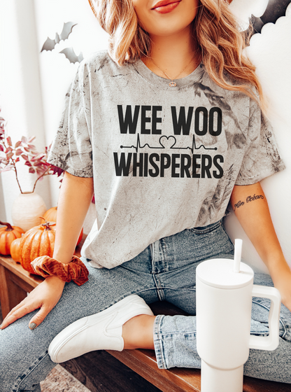 Person wearing a 'WEE WOO WHISPERERS' shirt sitting with a cup in a cozy indoor setting.
