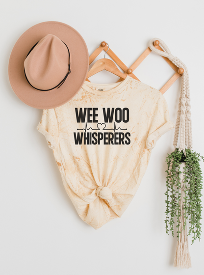 Beige tie-dye shirt with 'Wee Woo Whisperers' text, beige hat, wooden hangers, and plant on a white background